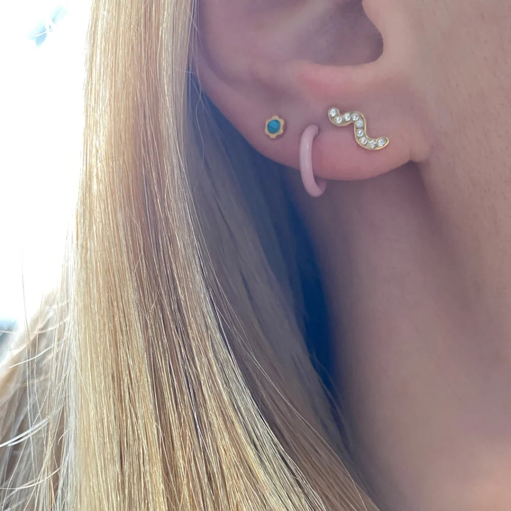 Ohr mit drei Ohringen: schlangeförmiger Stecker mit Diamanten, kleine Creole in lila und kleiner goldener Stecker mit grünem Stein.