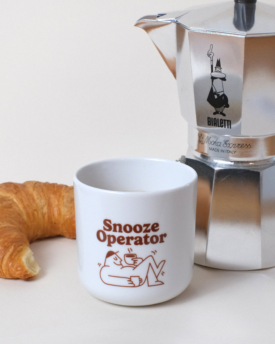 tasse mit braunem aufdruck snooze operator neben croissant und Bialetti Mokkakanne