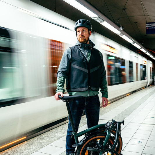 Mann mit schwarzer Fahrradwarnweste und Klapprad vor einfahrender Stadtbahn